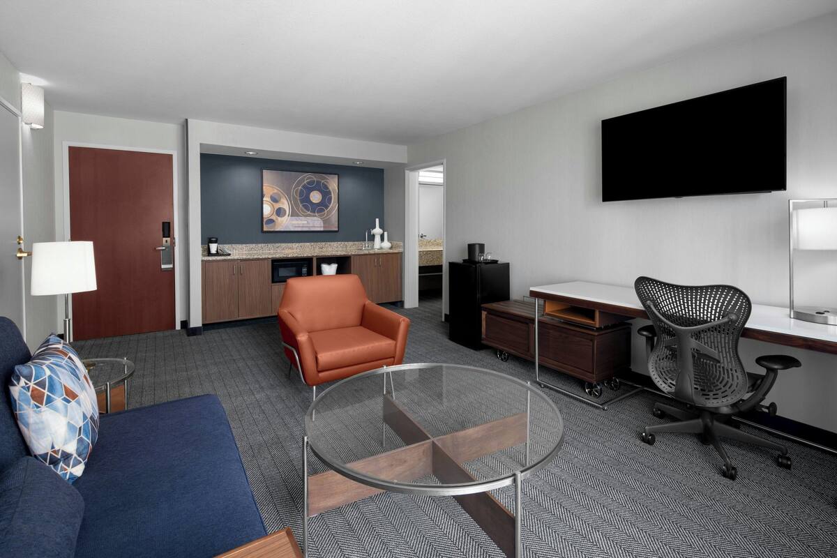 Ein modernes Hotelzimmer mit einer Couch, einem Glastisch, einem Schreibtisch mit Computer, einem Bürostuhl, einem Flachbildfernseher und einer Küchenzeile mit Schränken und einer Arbeitsplatte.