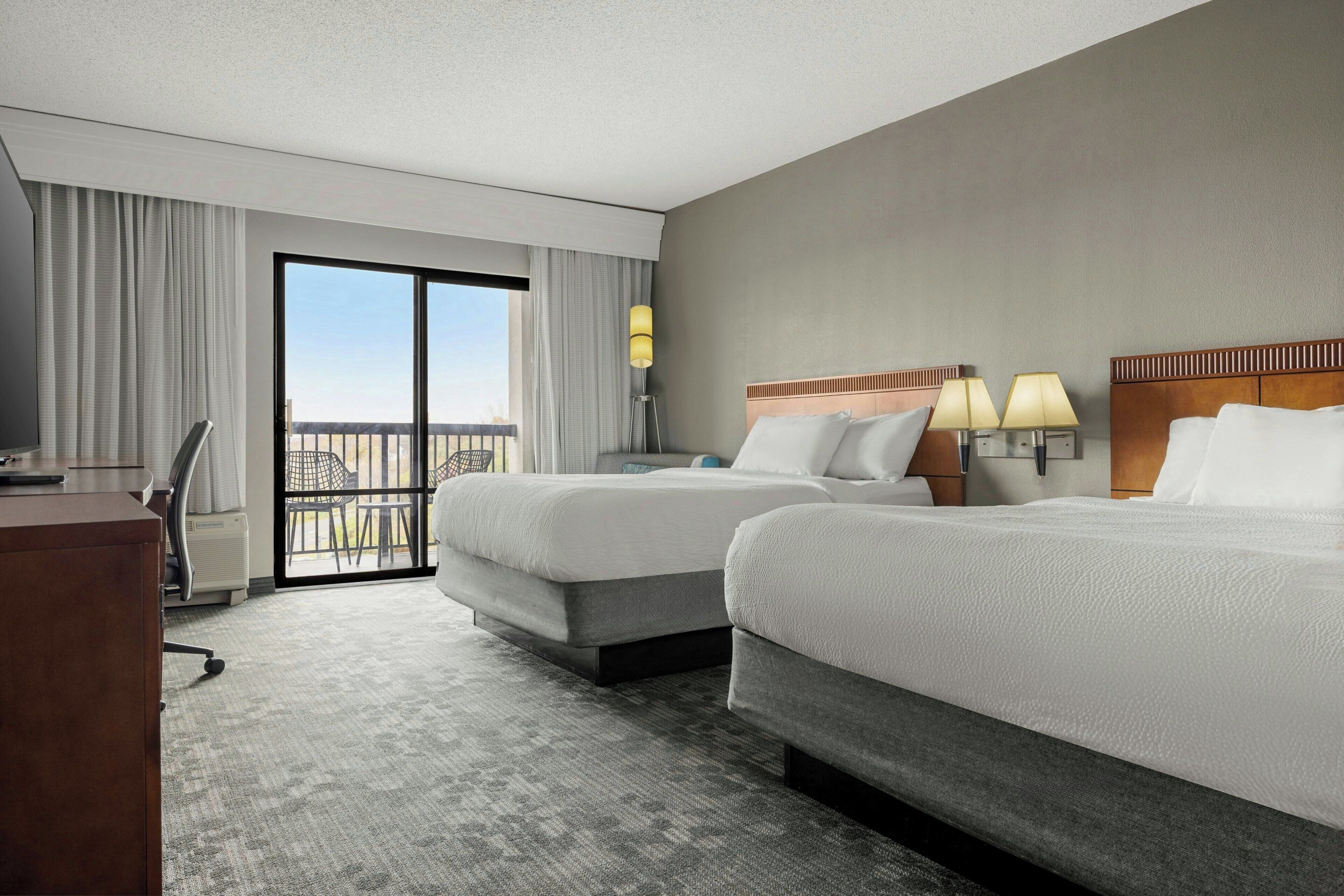 A hotel room with two beds, a desk, a chair, a lamp, and a view of the outdoors.