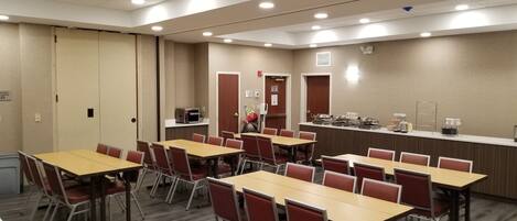 Un salón de conferencias con mesas y sillas, una kitchenette y una puerta con un cartel.