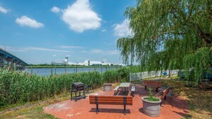 Een park aan het meer met een bank, picknicktafel, barbecue en potplanten.