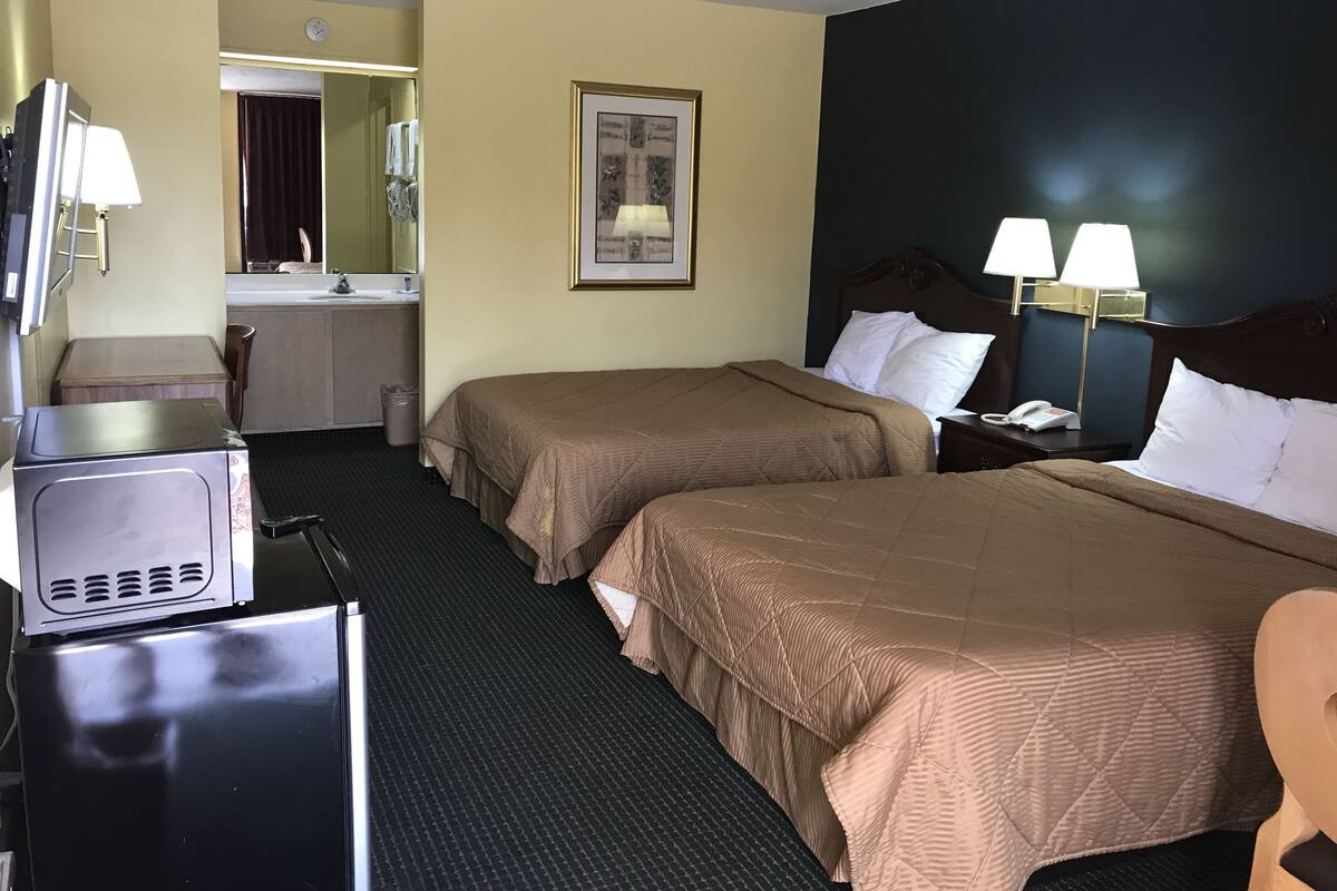 Habitación de hotel con dos camas, un escritorio con lámpara, un espejo, un cuadro en la pared y un cubo de basura.