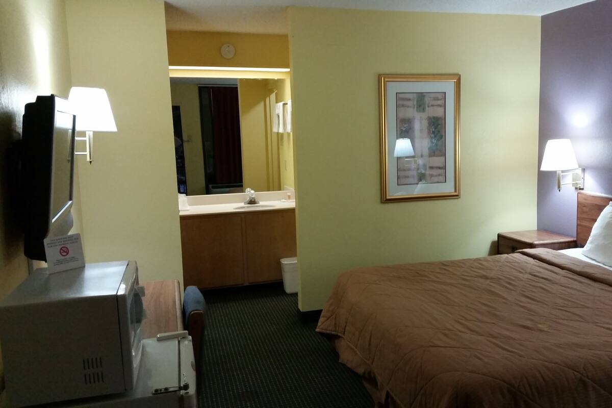Una habitación de hotel con cama, escritorio con lámpara, baño con lavabo y espejo, y un cuadro en la pared.