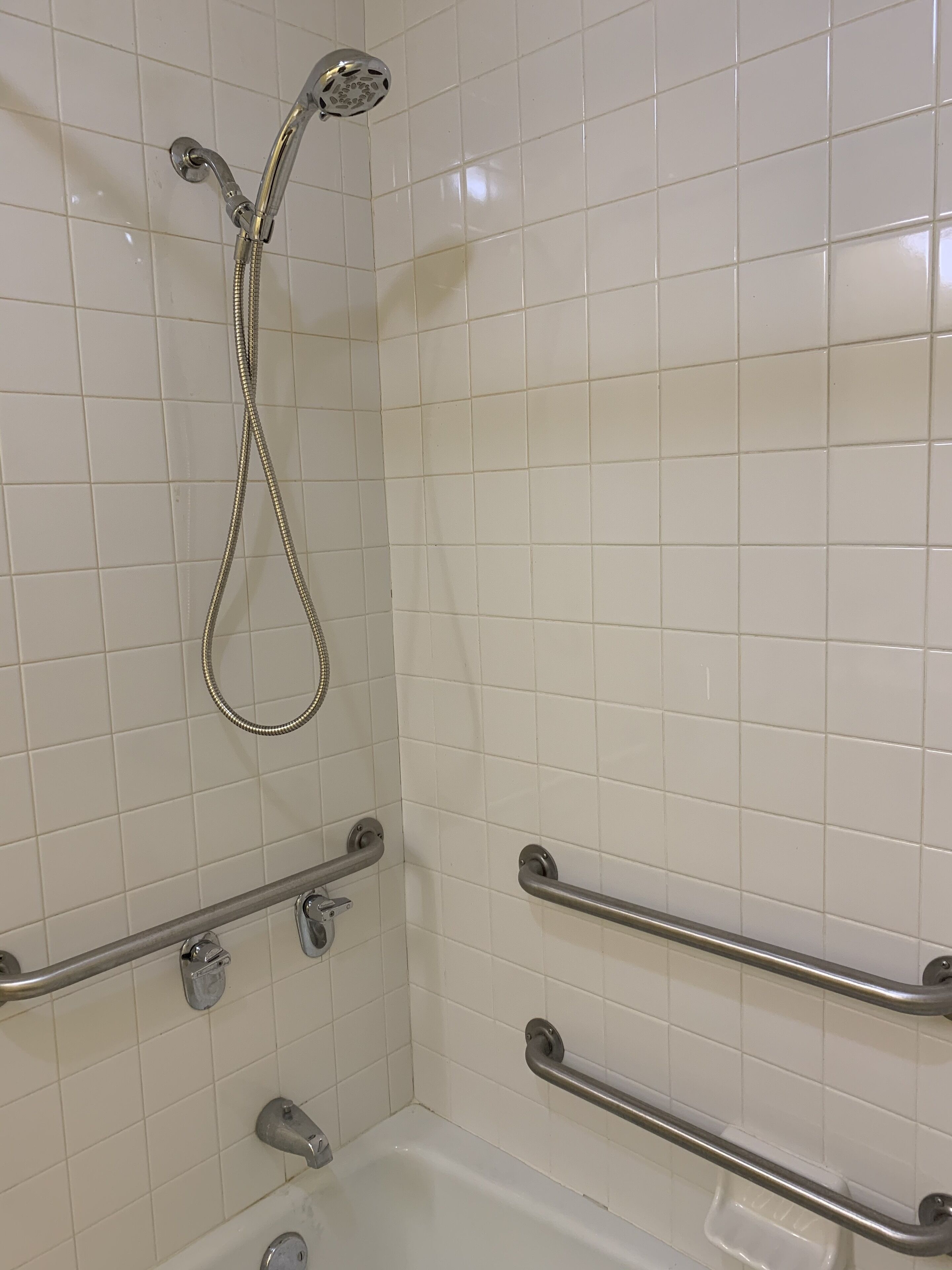 A bathroom with white tiled walls, a bathtub with a handheld showerhead, and two grab bars installed for safety.