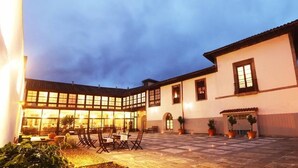 A courtyard with a dining area, potted plants, and a building with large windows and a tiled roof. - Hotel PAX Casona del Busto (Pravia)