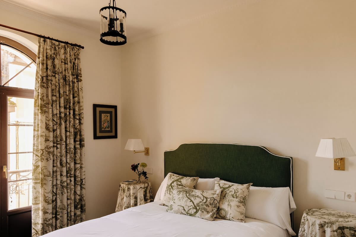 Uma cama bem arrumada com cabeceira verde, duas mesas de cabeceira com abajures, uma janela com cortinas florais e um quadro fixado na parede.