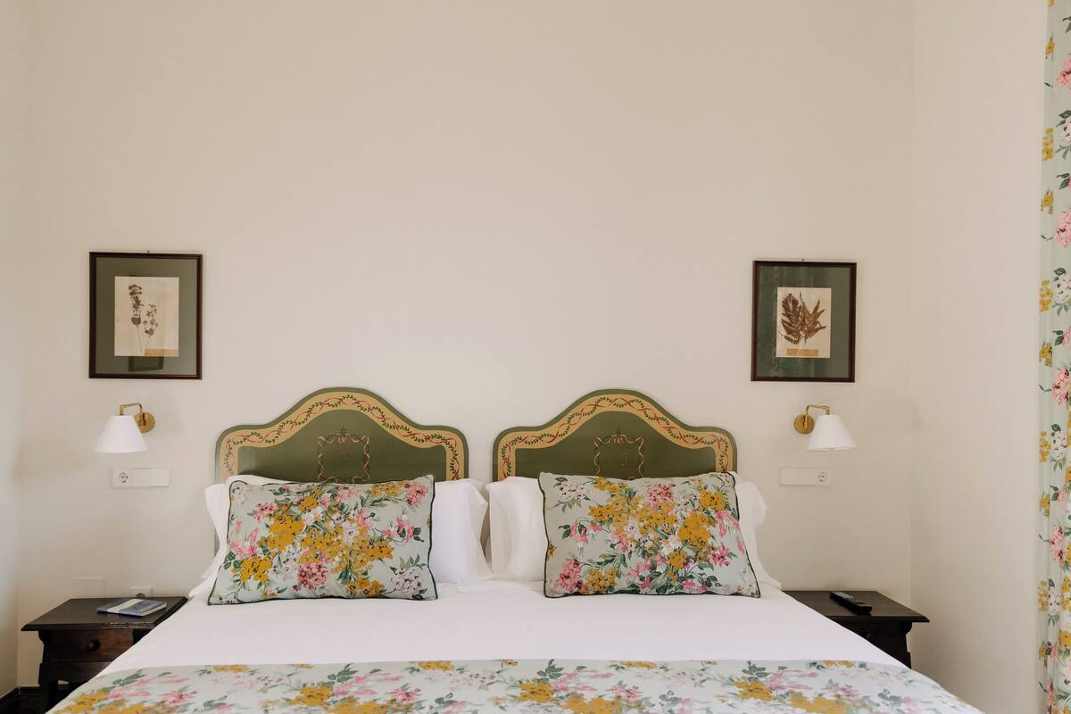 Uma cama bem arrumada com roupa de cama floral, duas mesas de cabeceira com abajures e arte botânica emoldurada nas paredes.