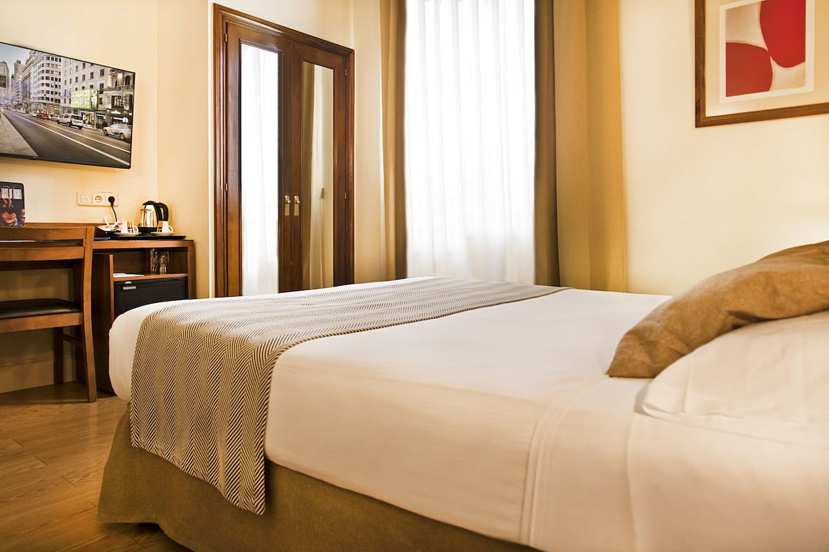 A hotel room with a bed, a desk, a chair, a small table with a kettle, and a framed picture on the wall.