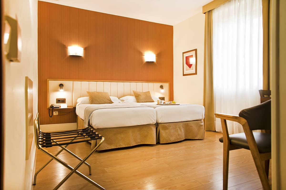 A hotel room with a double bed, two bedside lamps, a wooden headboard, a wooden floor, a chair, and a small table with a tray.