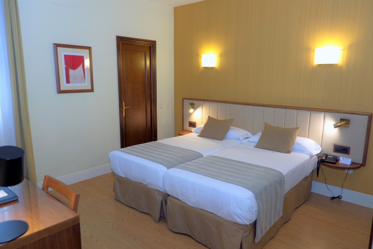 Habitación de hotel con dos camas, un escritorio, una silla, una puerta y dos luces fijadas a la pared.