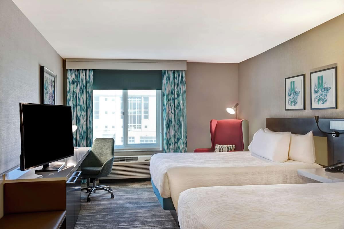 A hotel room with a bed, a desk with a TV, a chair, a desk lamp, and a window with curtains.