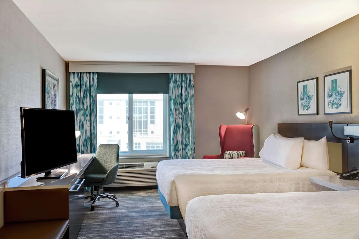 A hotel room with a bed, a desk with a computer, a TV, a chair, and a window with curtains.