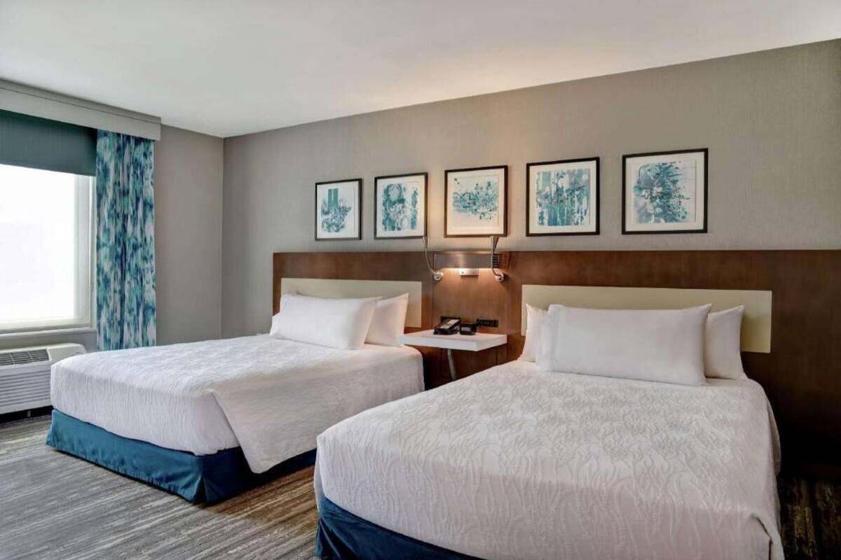 A hotel room with two beds, a headboard, a bedside table with a lamp, and framed artwork on the wall.