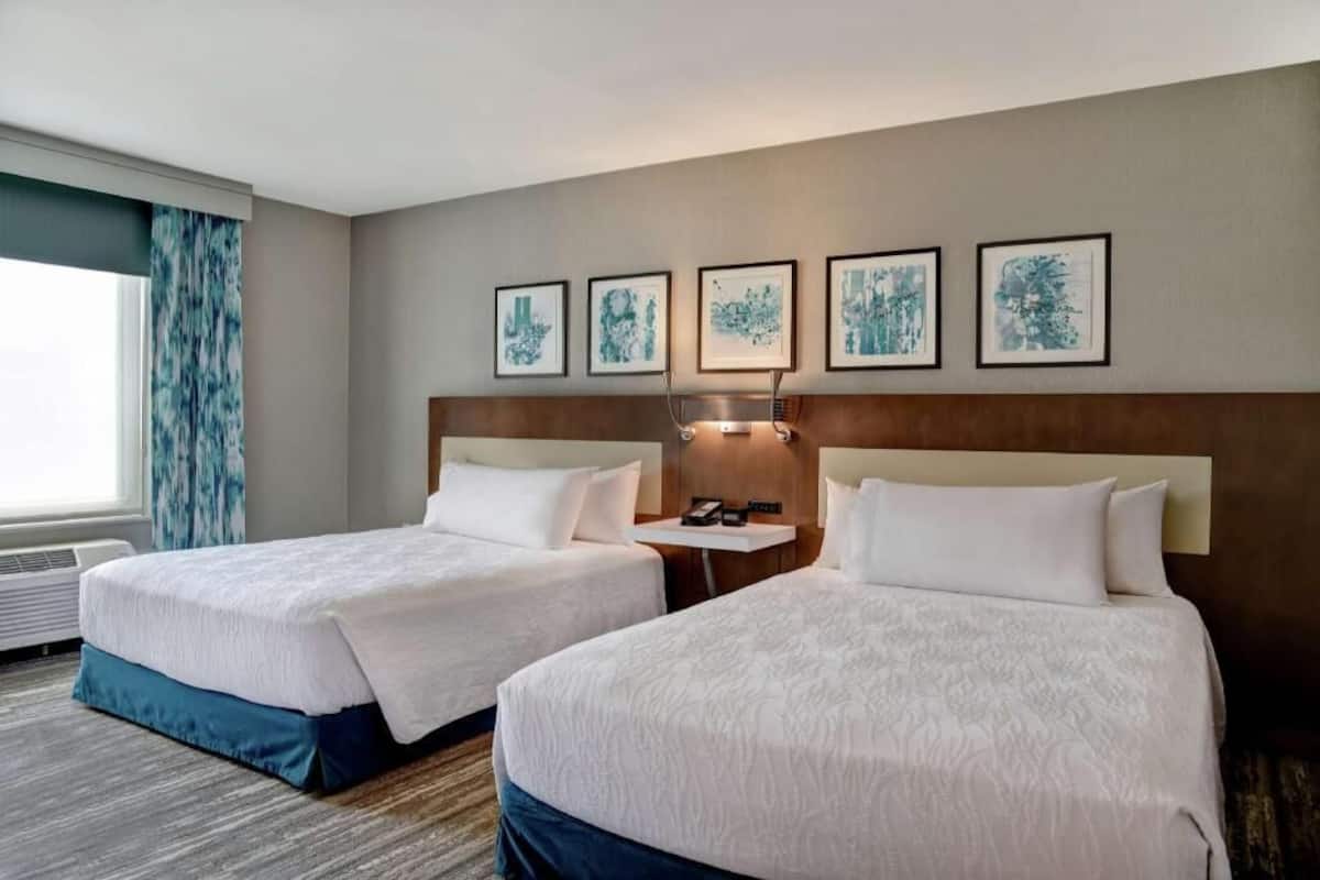 A hotel room with two beds, a headboard, a bedside table with a lamp, and framed artwork on the wall.