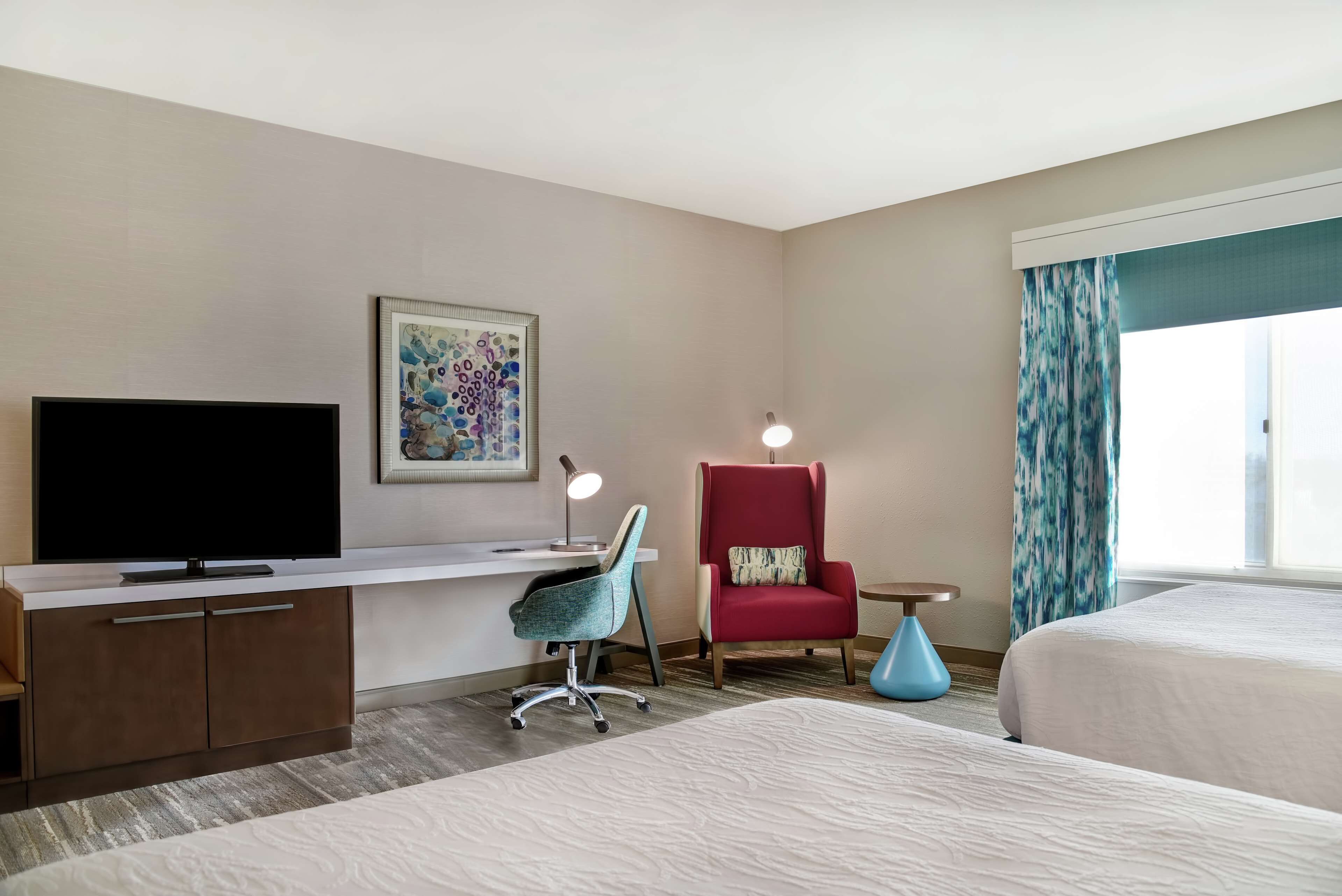 Quarto de hotel com uma cama, uma escrivaninha com televisão, uma cadeira vermelha, uma cadeira azul e um quadro na parede.