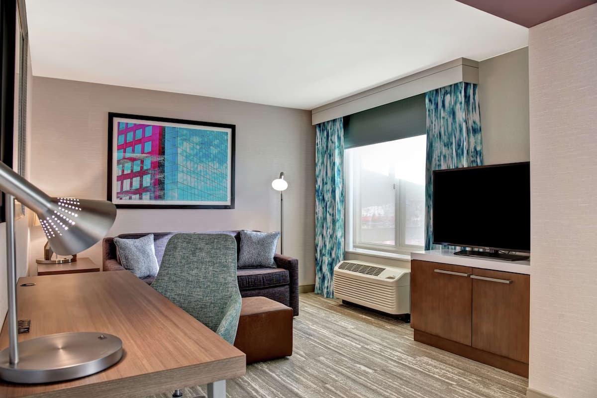 A modern hotel room with a sofa, a desk with a lamp, a flat-screen TV, and a large window with curtains.