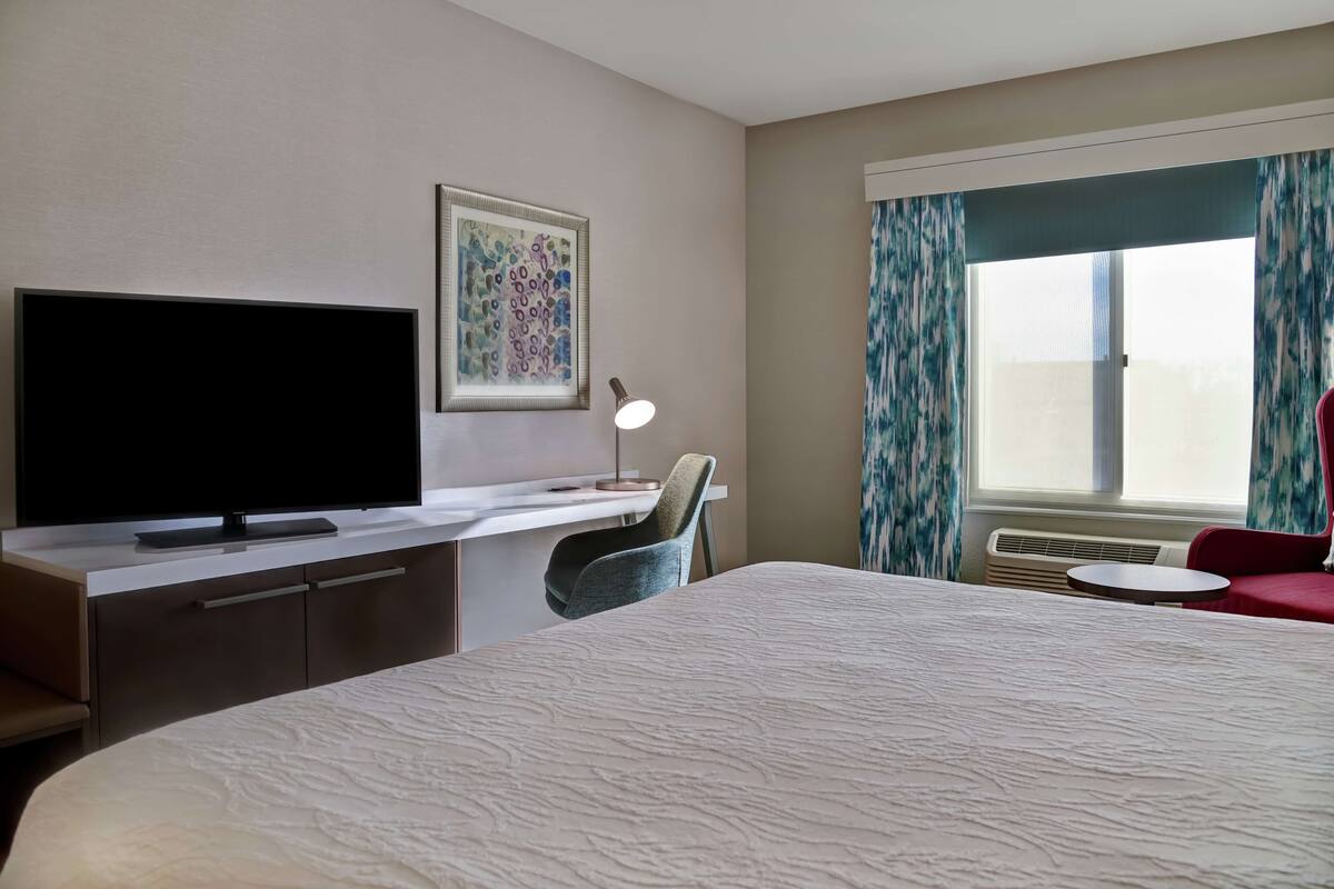 A hotel room with a bed, a desk with a flat-screen TV, a chair, a desk lamp, a window with curtains, and a wall-mounted artwork.