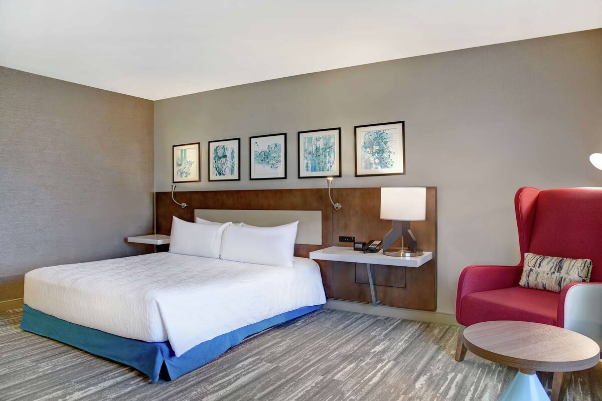 A hotel room with a large bed, a red armchair, a small round table, and a bedside table with a lamp. There are four framed artworks on the wall above the bed.