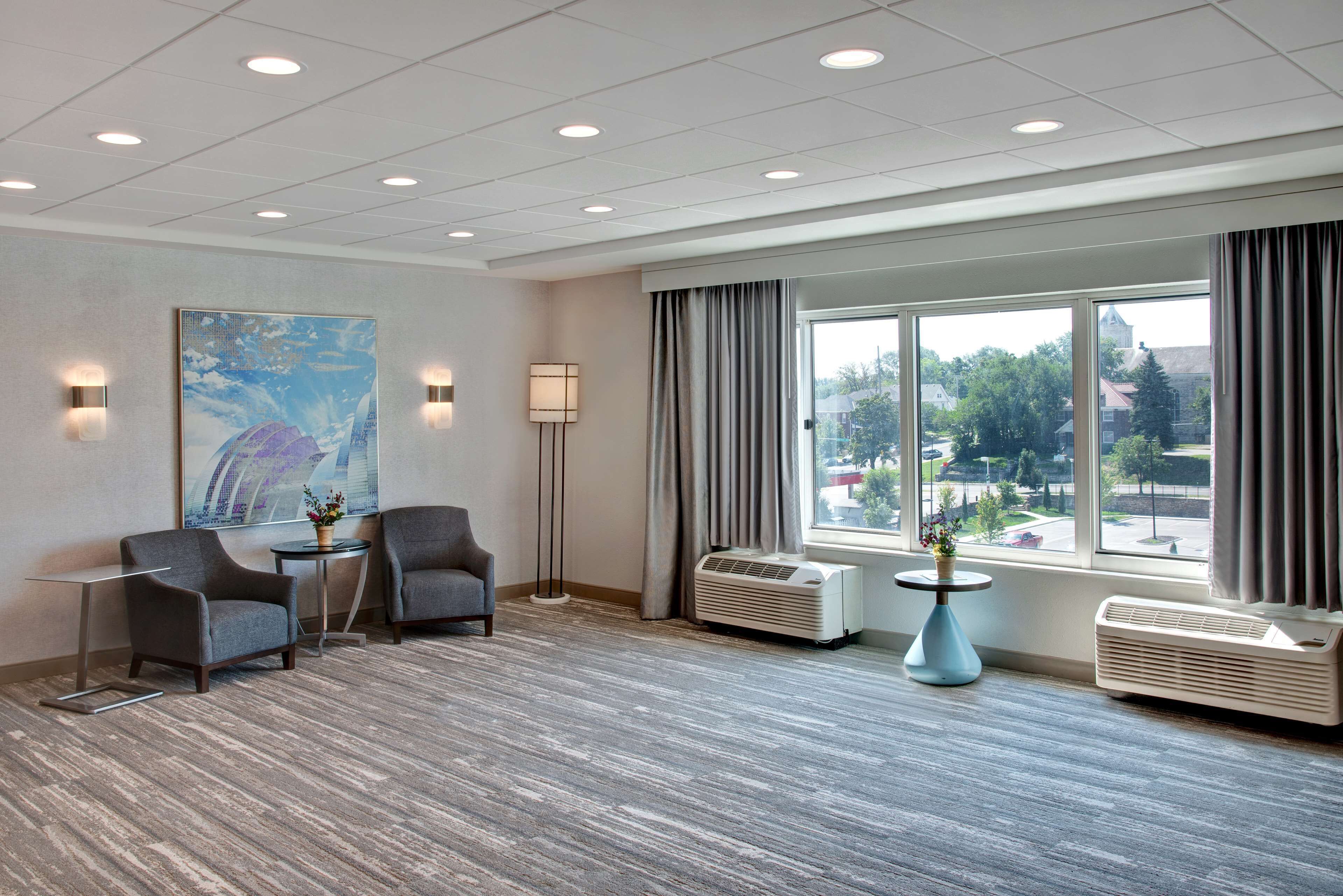 A hotel room with two armchairs, a small table, a large window with curtains, an air conditioning unit, and a painting on the wall.
