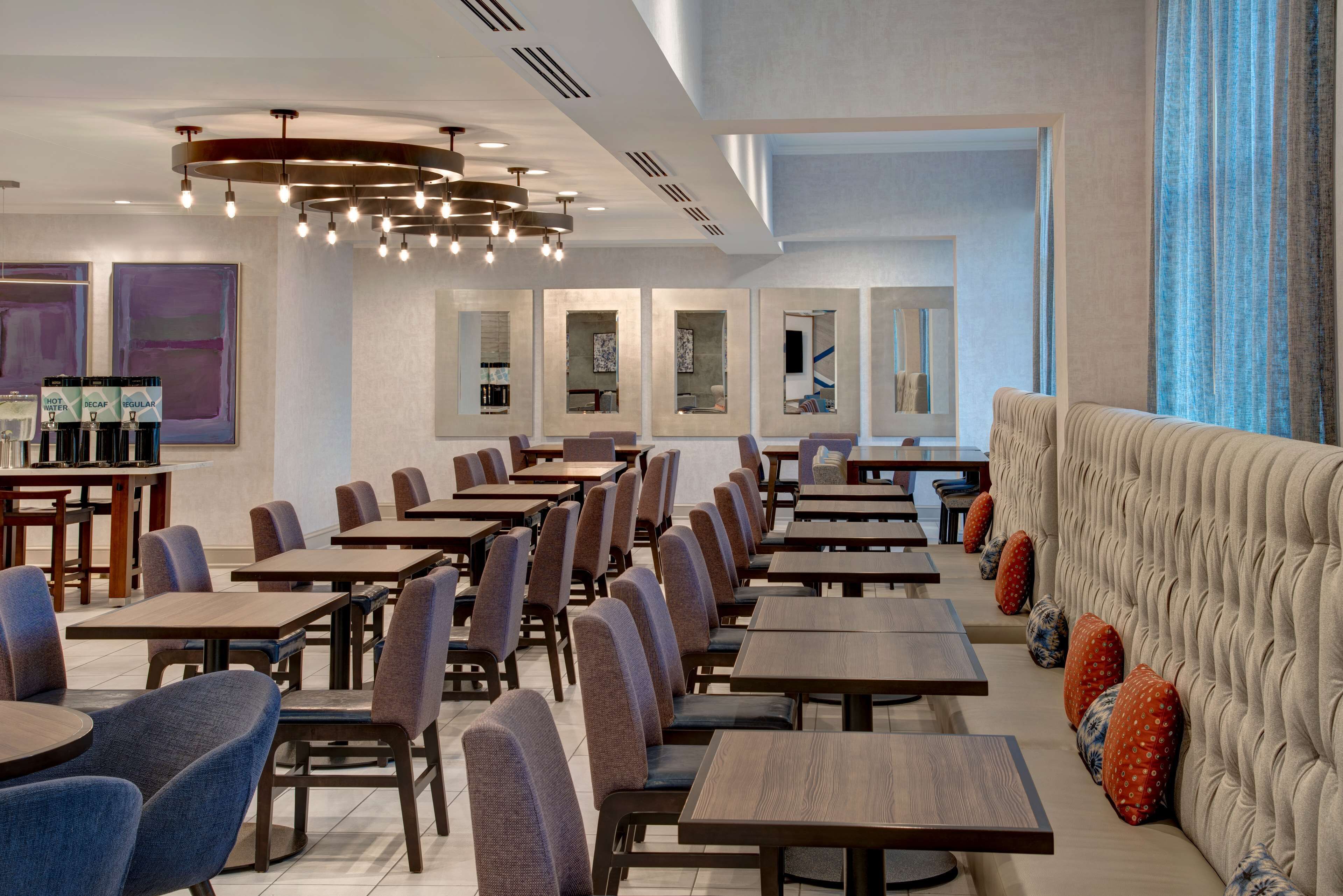 Uma sala de jantar moderna com várias mesas e cadeiras, um sofá com almofadas decorativas e um teto com luminária única.