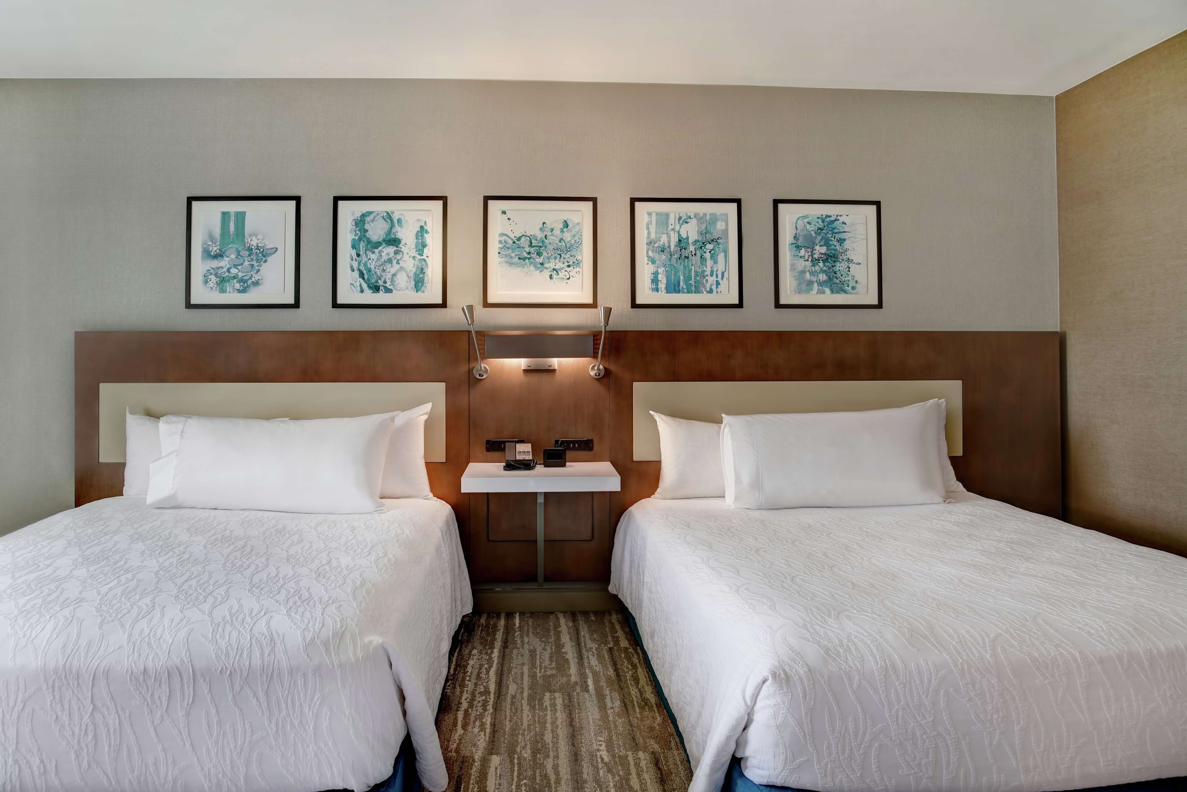 Una habitación de hotel con dos camas, una mesita pequeña y obras de arte en la pared.