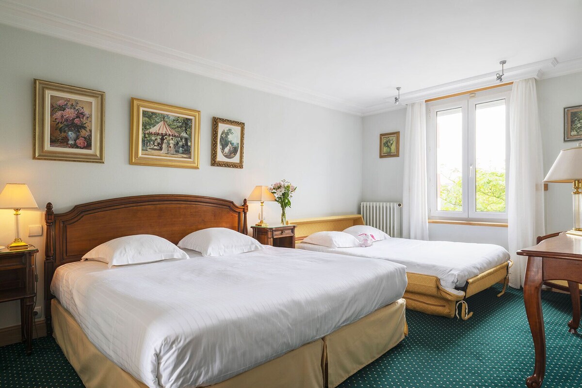 A hotel room with two beds, a wooden headboard, a radiator, a window with curtains, and framed artwork on the wall.