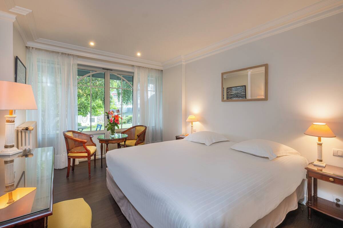 A hotel room with a large bed, two bedside lamps, a round table with chairs, a mirror on the wall, and a window with curtains.