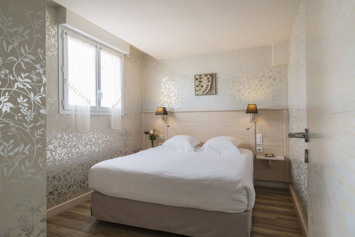 A bedroom with a bed, two bedside lamps, a nightstand, a wall clock, and a window with sheer curtains.