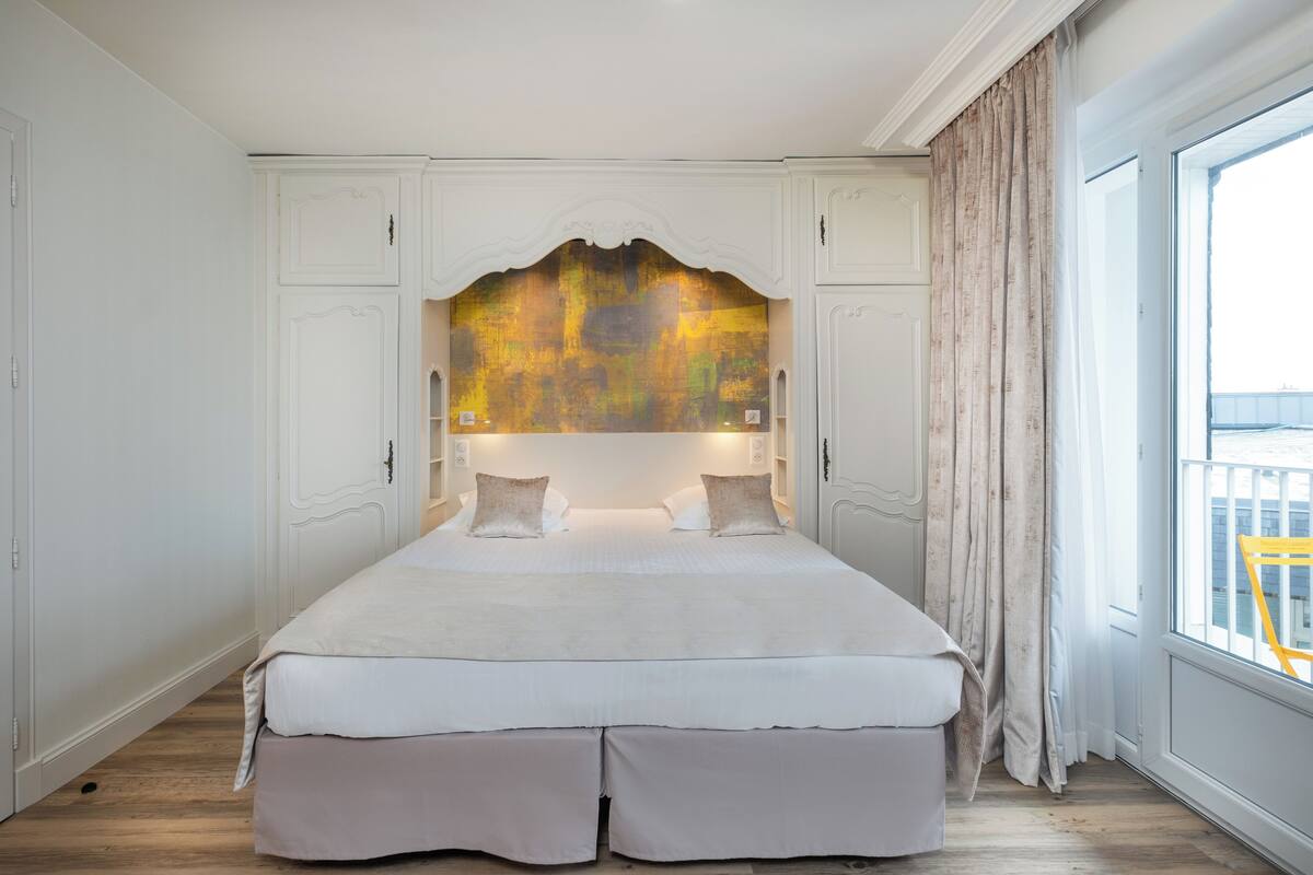 A large bed with a decorative headboard, white walls, and a view of the outside through a window.
