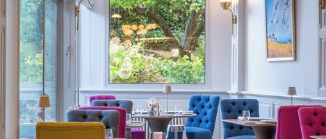 Ein Restaurantinneres mit einem großen Fenster, das einen Blick auf Grünflächen und einen Teich bietet. Es gibt mehrere Esstische mit Stühlen, und der Raum hat einen klassischen architektonischen Stil mit weißen Wänden und dekorativer Beleuchtung.