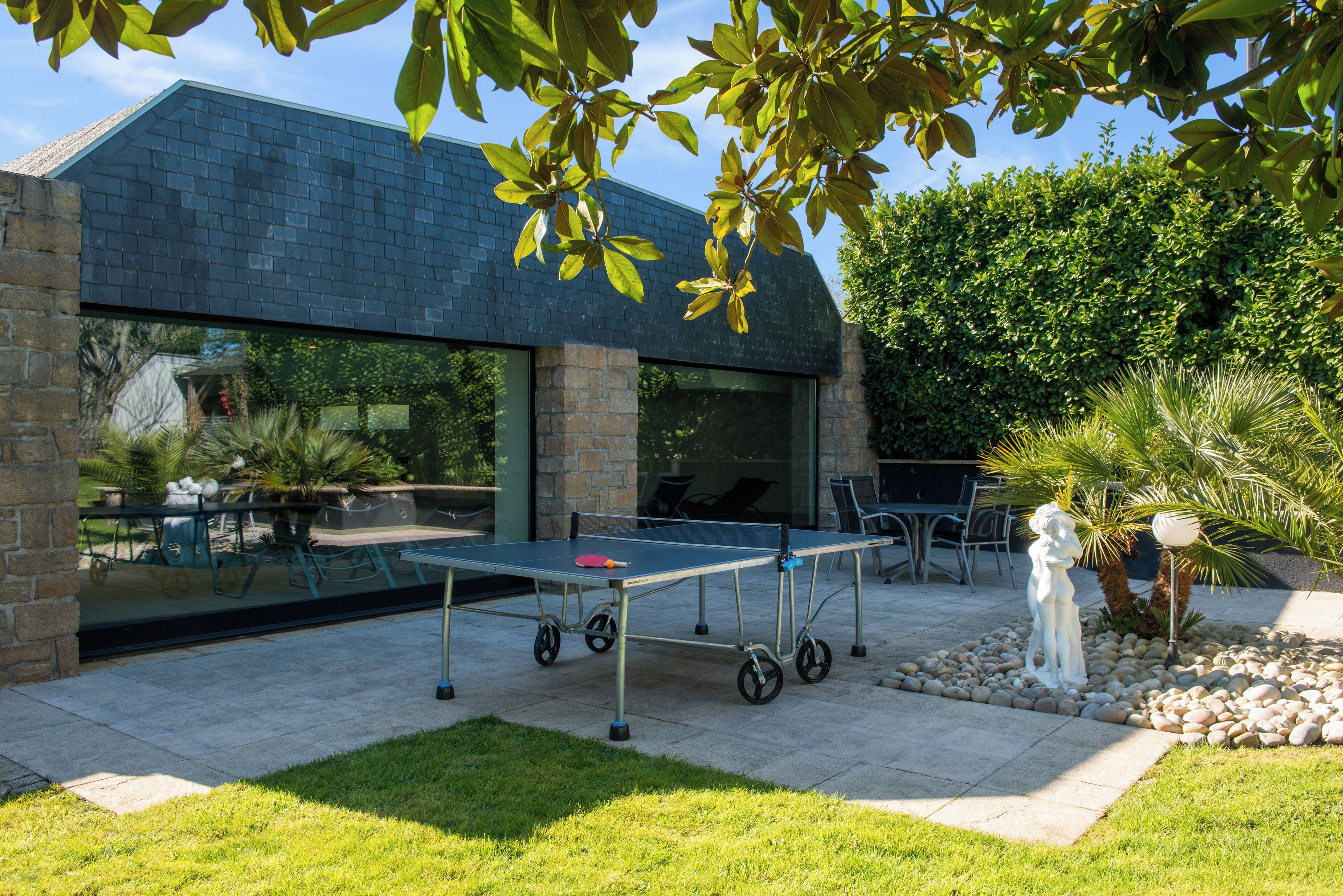 Une table de ping-pong est installée dans un espace patio, avec un mur en pierre et une porte vitrée donnant sur un espace de détente extérieur abrité.