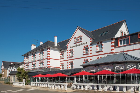 A hotel with a red and white facade, multiple windows, and outdoor seating with red umbrellas.