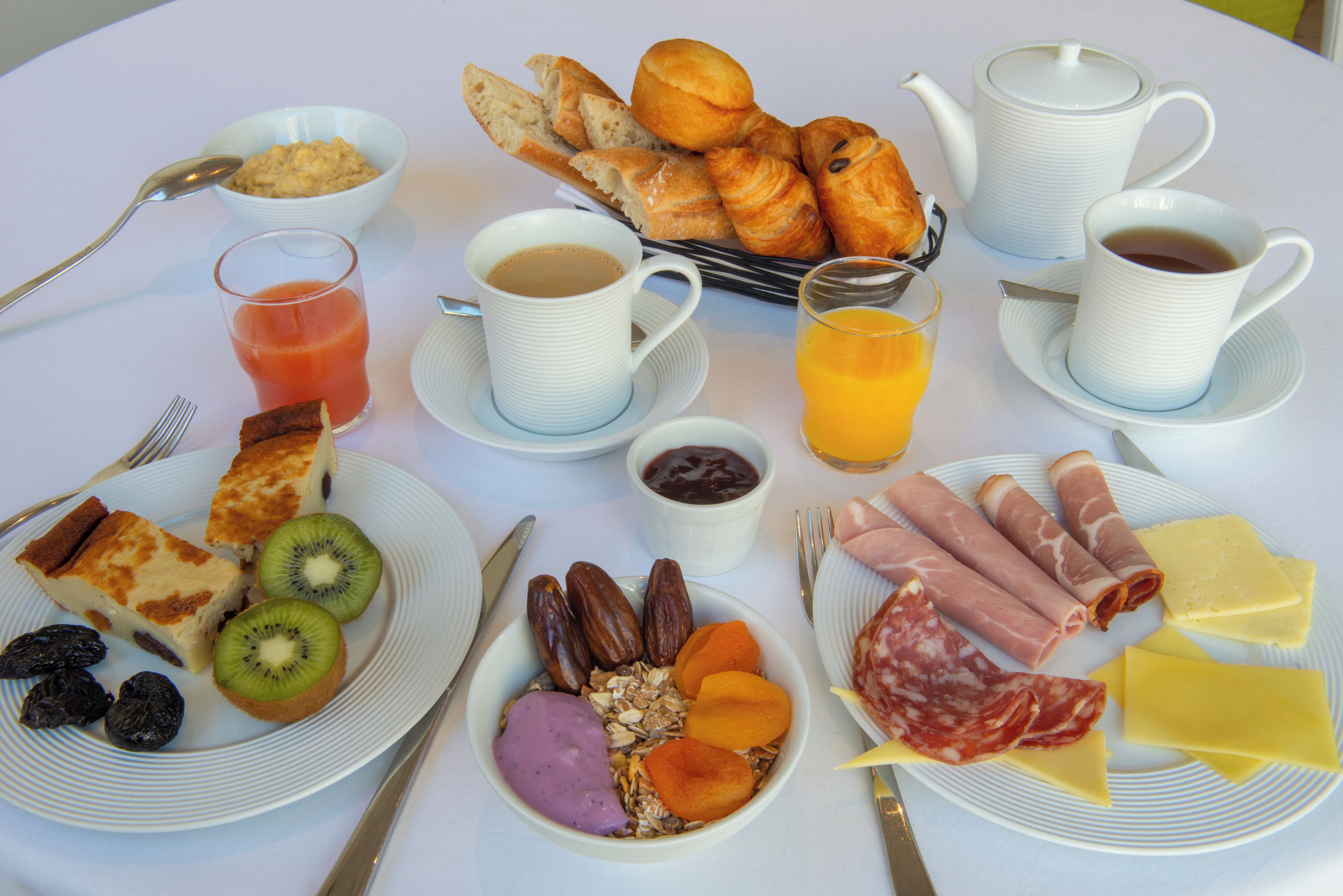 Una mesa de desayuno con cruasanes, café, jugo y una variedad de quesos y embutidos.