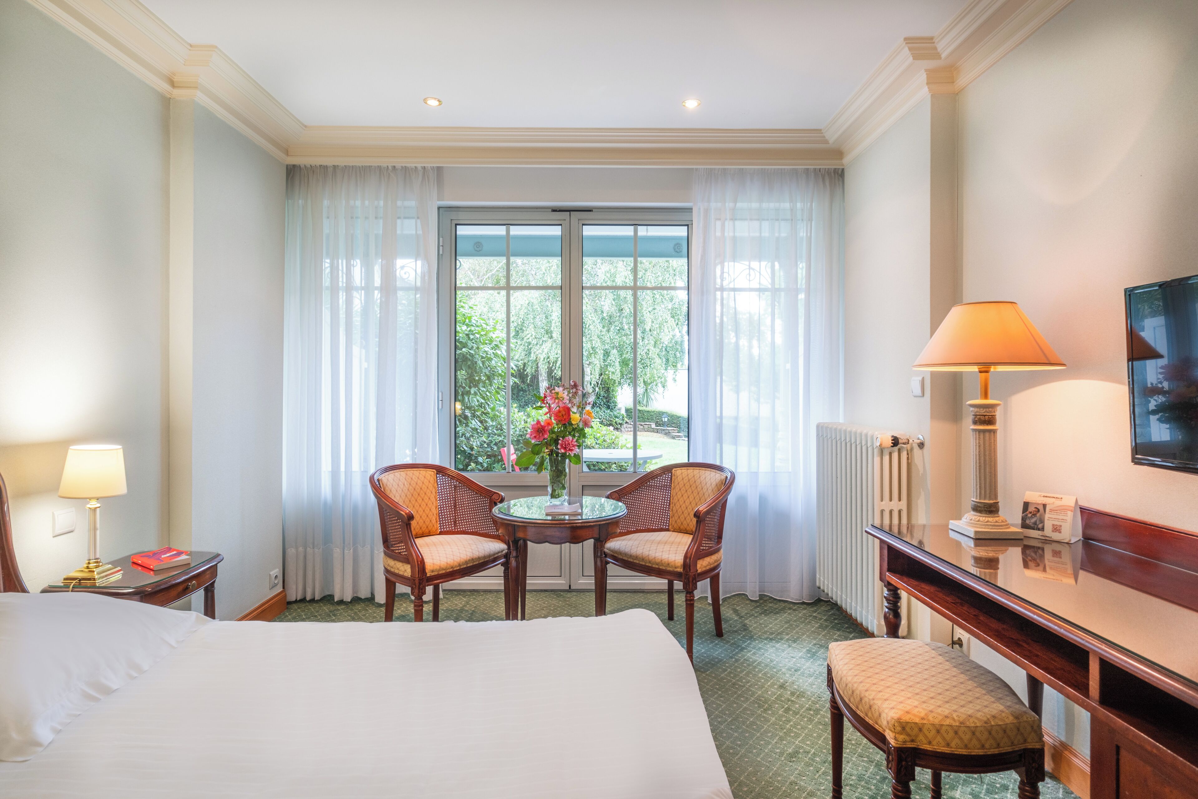 Une chambre d’hôtel comprenant un lit, deux chaises, une petite table avec un vase de fleurs, un bureau avec une lampe et une vue sur la verdure par la fenêtre.