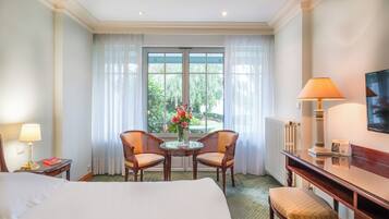 Une chambre d’hôtel comprenant un lit, deux chaises, une petite table avec un vase de fleurs, un bureau avec une lampe et une vue sur la verdure par la fenêtre.