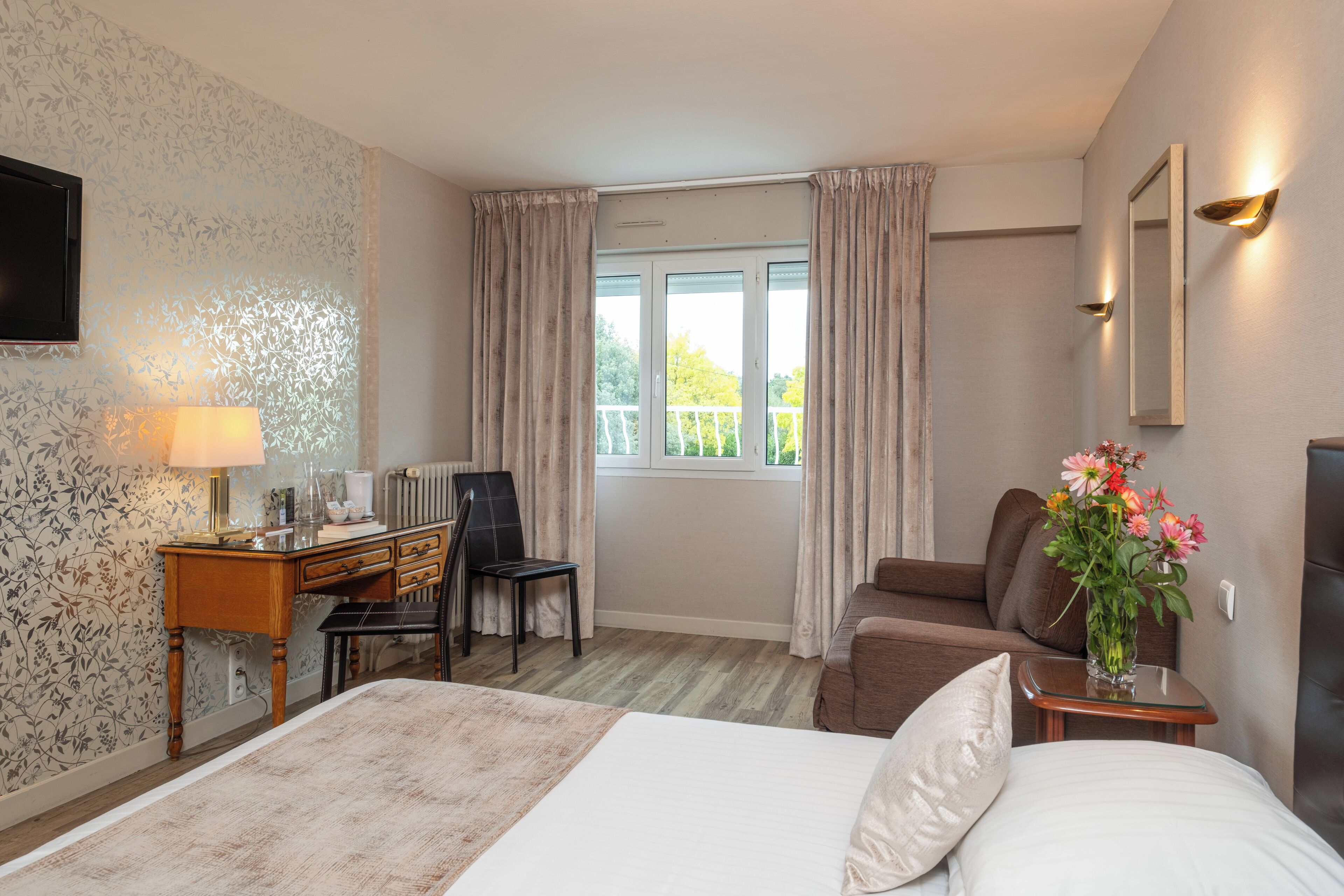 A hotel room with a bed, a desk, a chair, a sofa, a window with curtains, a vase with flowers, and a wall-mounted TV.