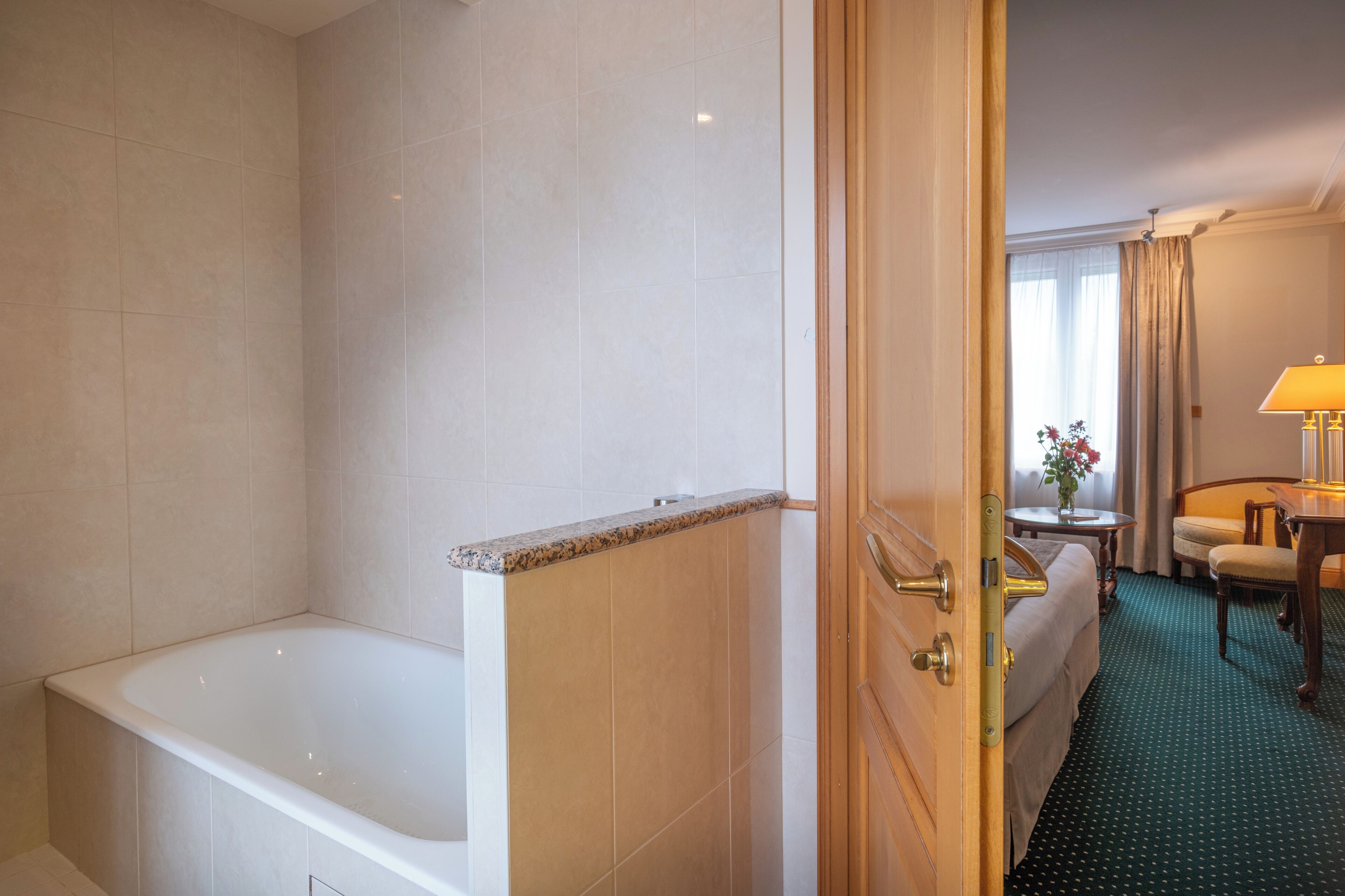 A hotel bathroom with a bathtub, a shower, and a toilet. There is a bed with a green carpeted floor and a small table with a lamp.