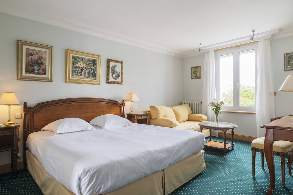 A hotel room with a large bed, two bedside tables, a chair, a small table with a vase, and a window with curtains.