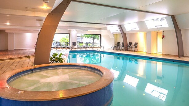 An indoor swimming pool with a hot tub, sunlit ceiling, and outdoor seating area visible through large windows. - Domaine du Limonay, The Originals Collection (Saint-Meloir-des-Ondes)