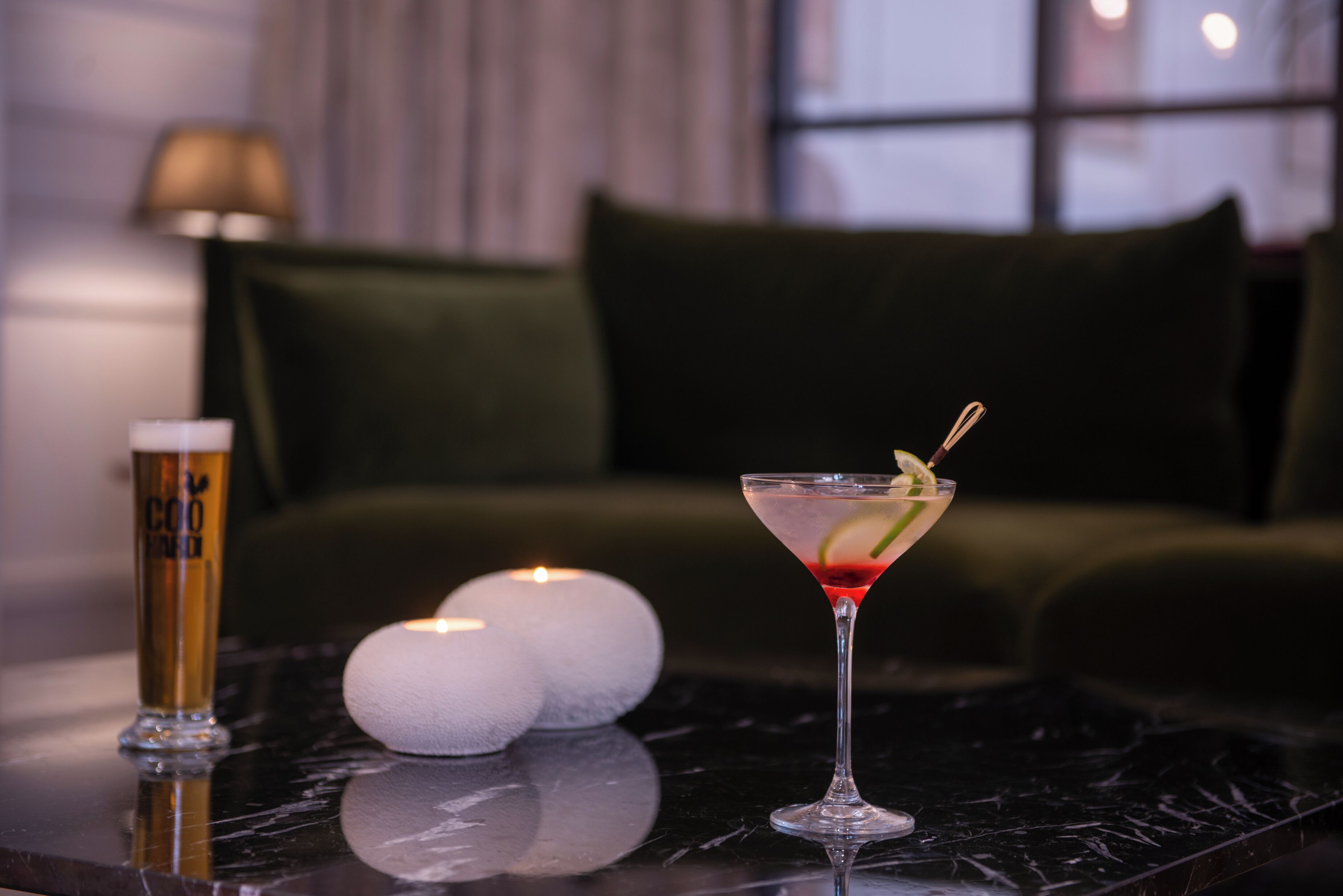 A martini glass with a lime garnish on a marble table, a tall beer glass, and two white candles in a dimly lit room with a green couch and a lamp in the background.