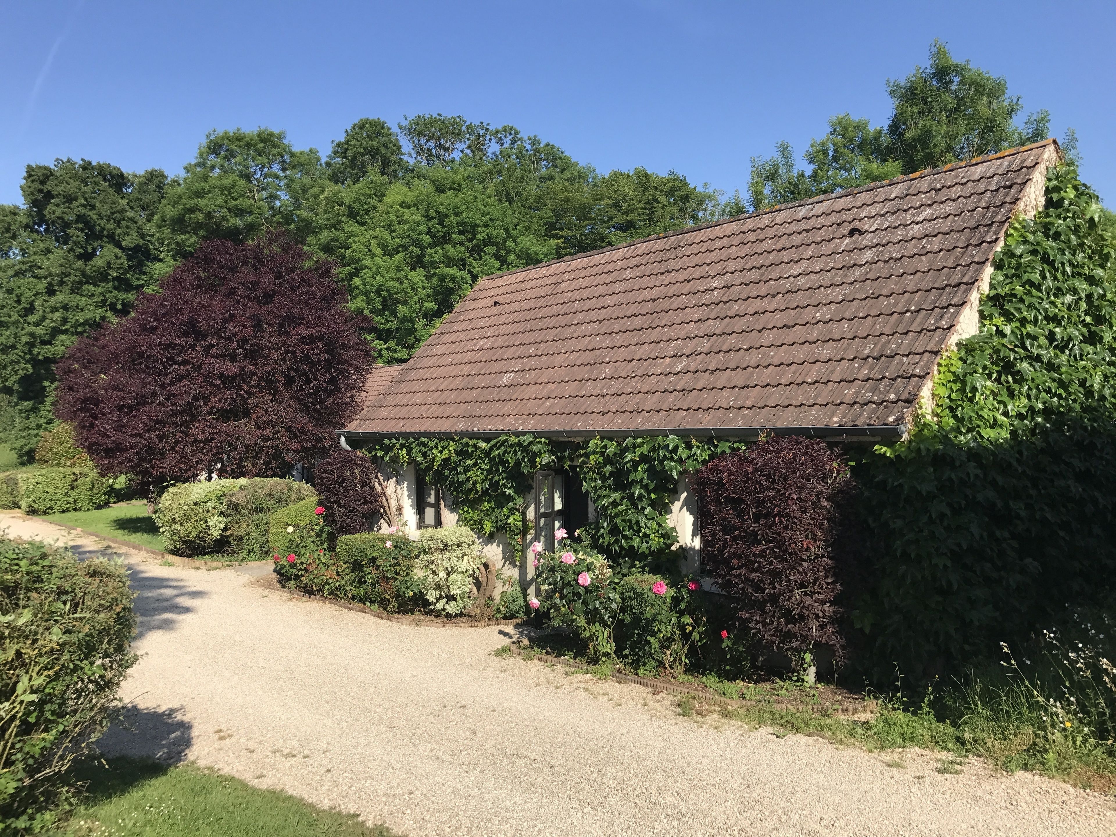 A rustic stone building with a sloped roof, surrounded by lush greenery and flowering plants.