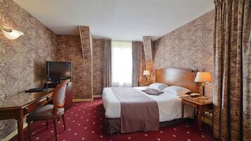 Une chambre d’hôtel avec un lit, un bureau avec une télévision, une chaise, une lampe et des rideaux.