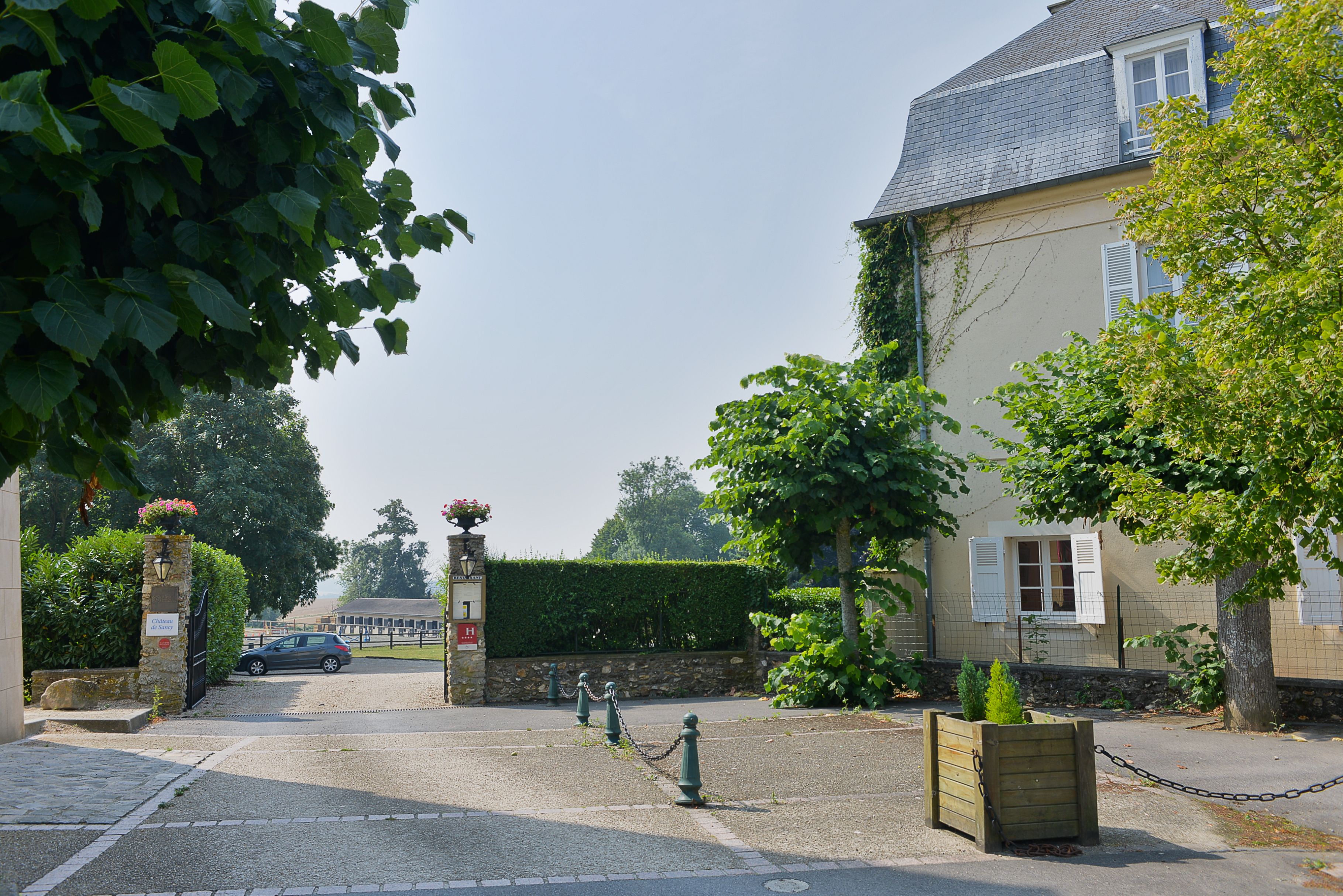 A gated entrance with a car parked inside, a stone wall, and a building with a sloped roof and shutters.