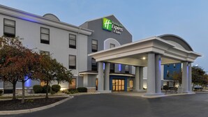 Edifício da Holiday Inn Express & Suites com fachada moderna, grande cobertura de entrada e área externa bem cuidada com árvores e um banco.