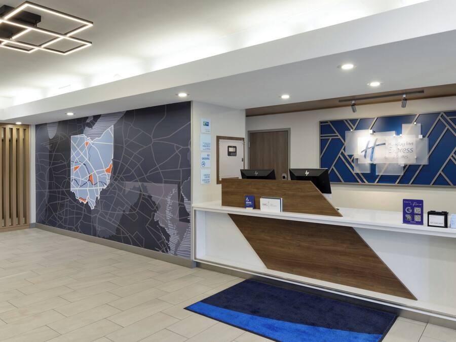 Uma área de recepção de hotel com design moderno, apresentando um balcão de atendimento, um mural com mapa e uma placa da Holiday Inn Express Suites.