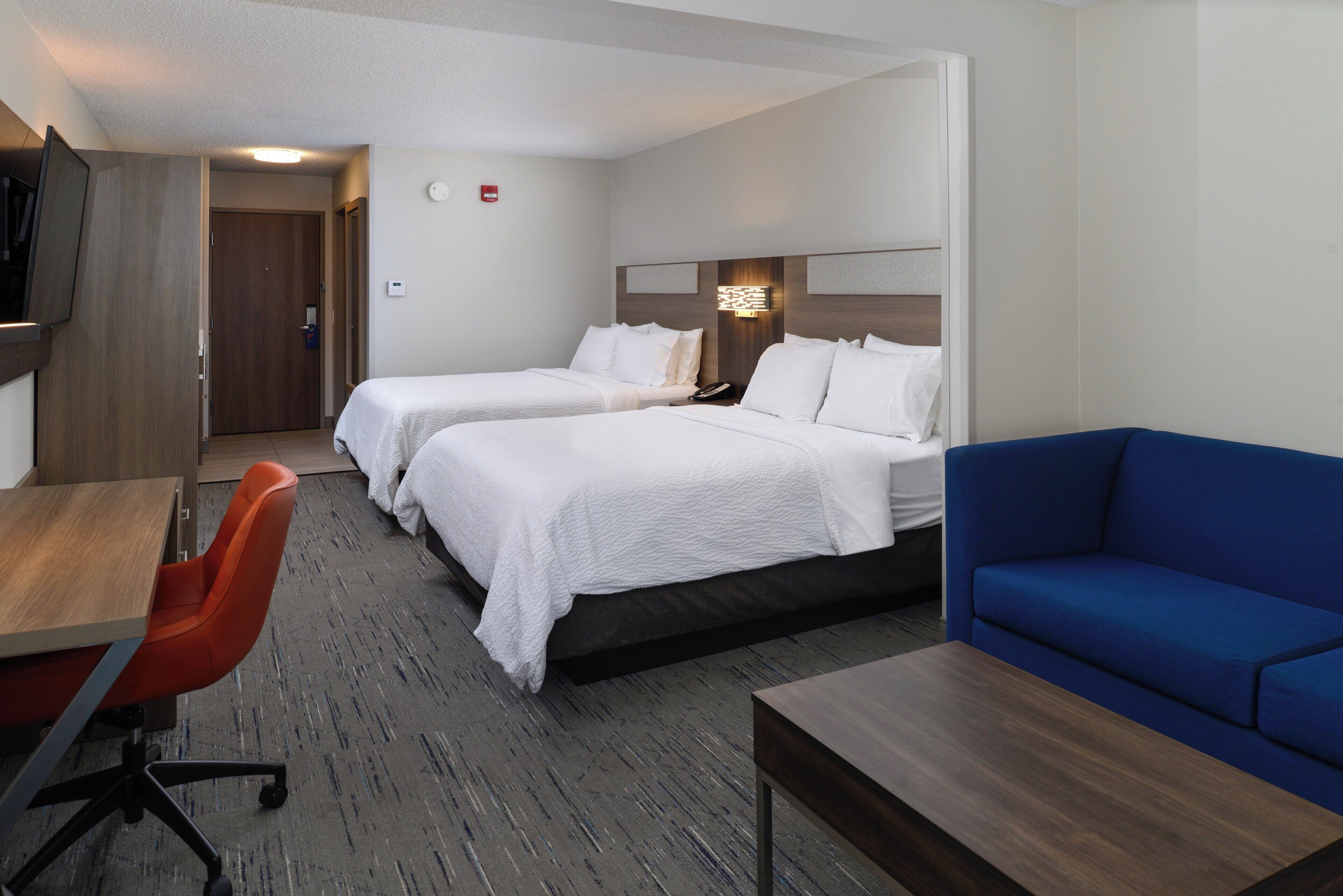 Una habitación de hotel con dos camas, un escritorio, un sofá azul y un televisor montado en la pared.