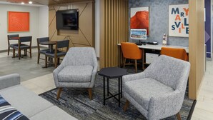 Um saguão de hotel moderno com poltronas revestidas de cinza, uma pequena mesa preta e uma televisão fixada na parede.
