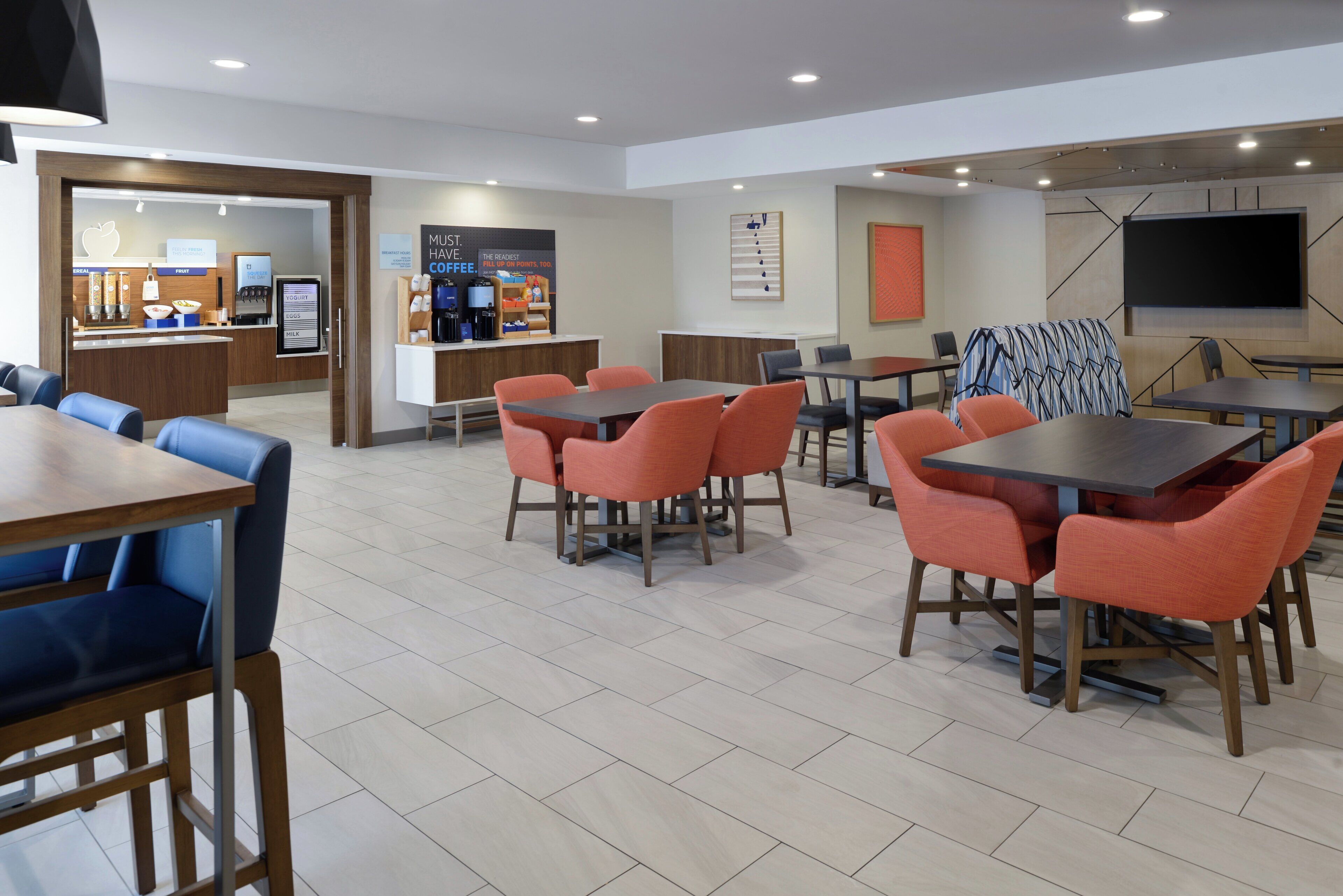 A modern hotel lobby with a coffee bar, seating area with tables and chairs, and a large flat-screen TV mounted on the wall.