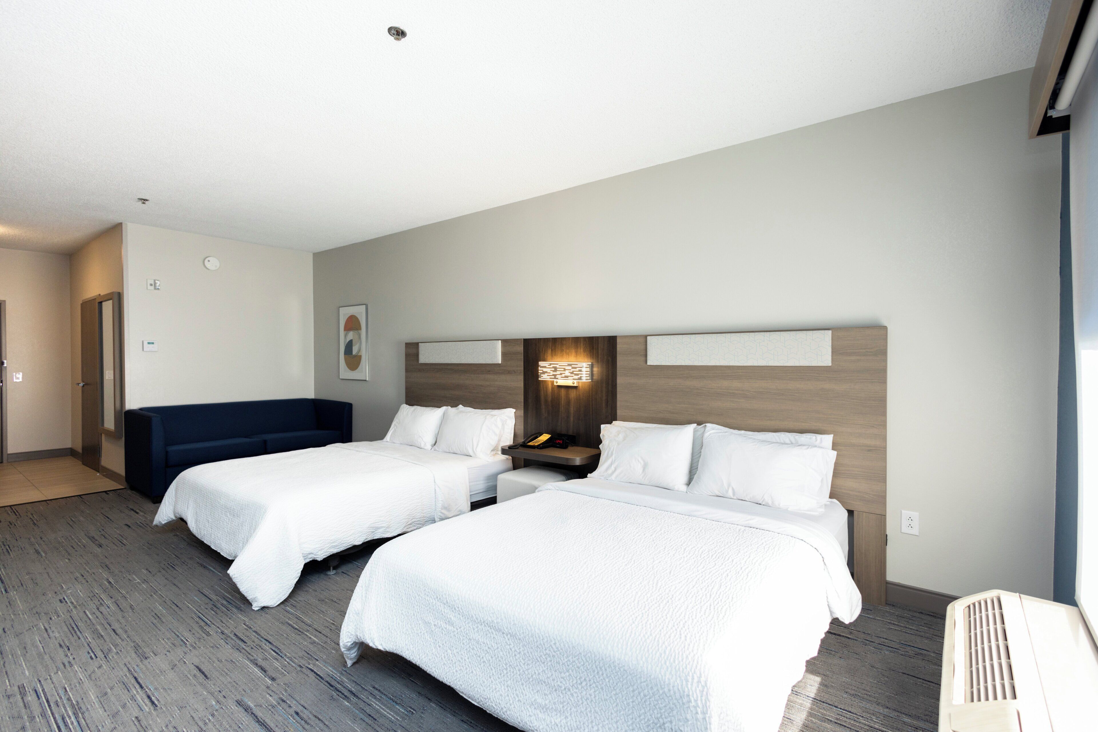 A hotel room with two beds, a sofa, a wall-mounted light, and a large window.