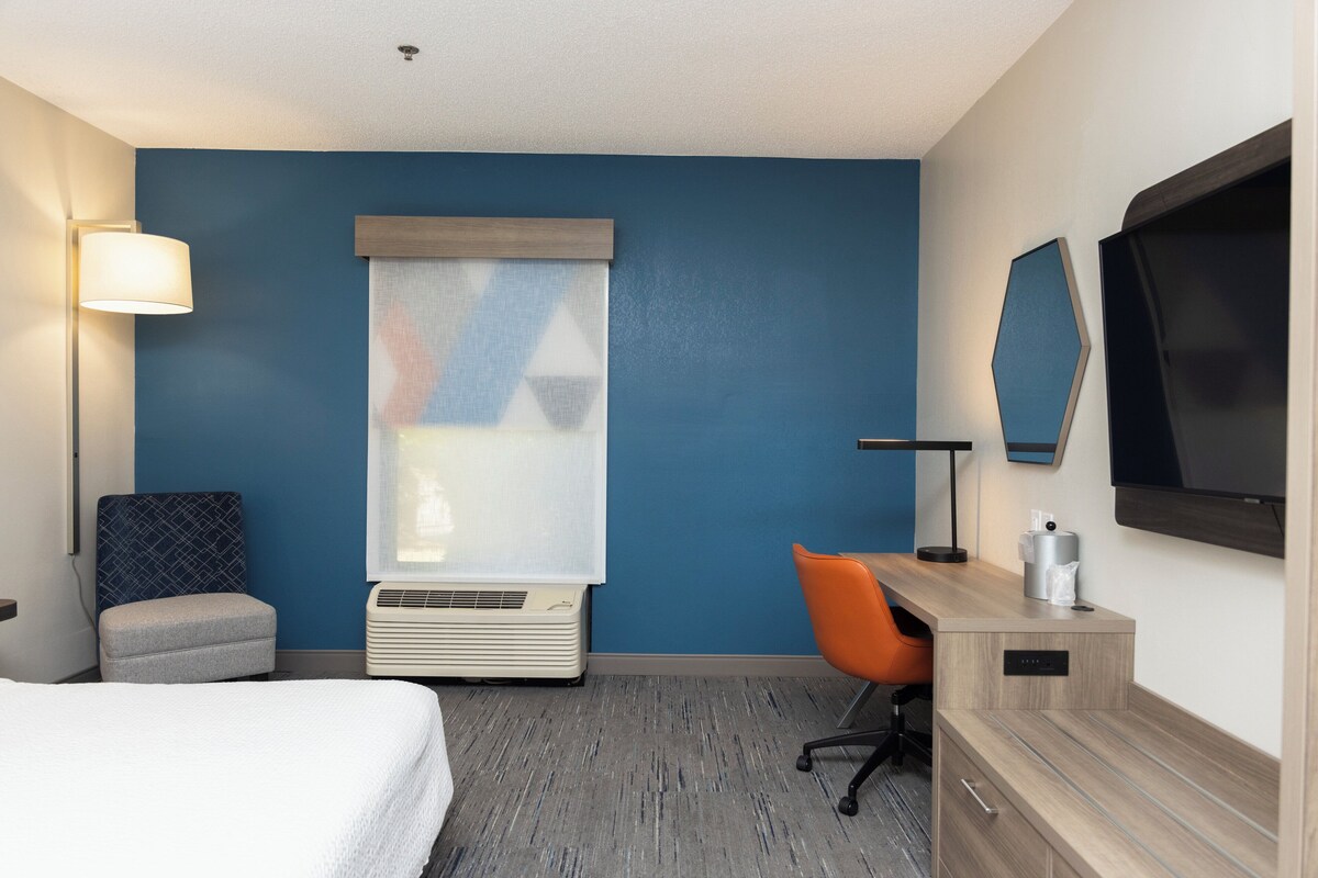 A hotel room with a bed, a desk with an orange chair, a TV, a lamp, and a window with blinds.