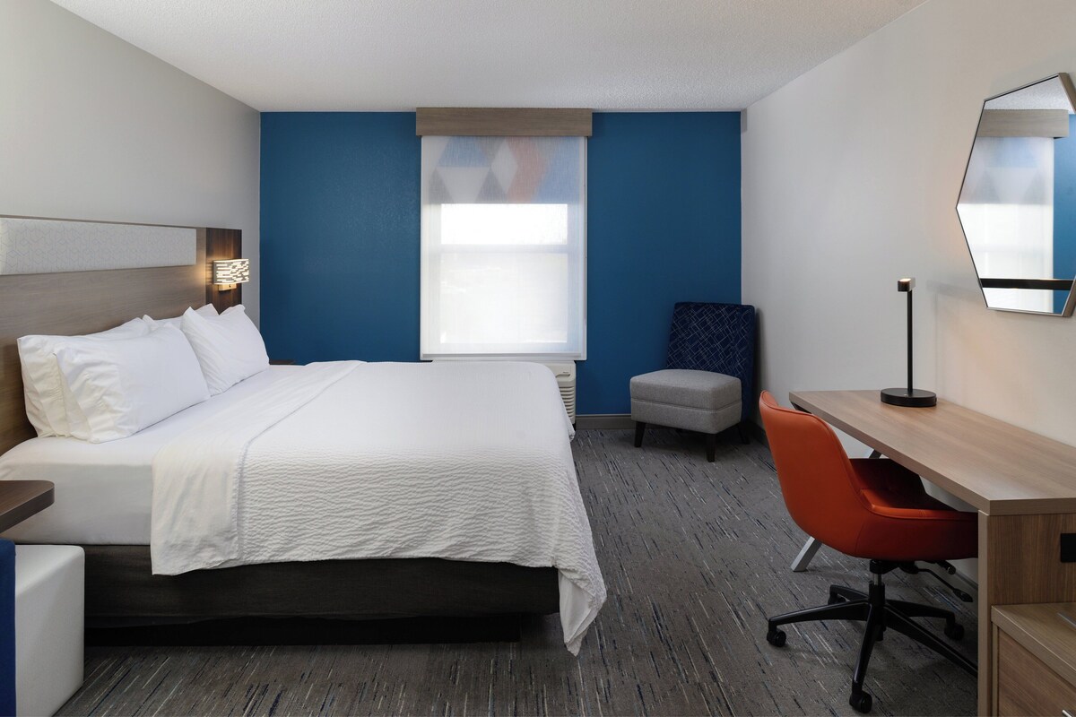 A hotel room with a bed, a desk, an orange chair, a large mirror, and a window with blinds.